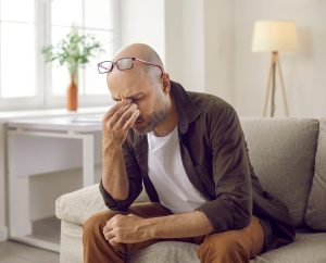 Man sitting indoors and pinching the bridge of his nose due to headache discomfort.